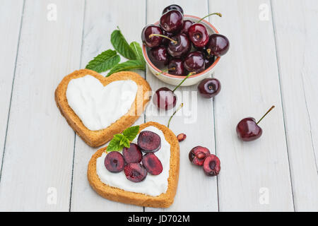 Schüssel gefüllt mit Kirschen und herzförmige Kekse mit Quark, Kirschen und einem Zweig Minze zu verbreiten Stockfoto