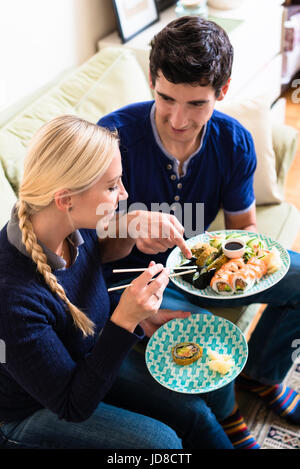 Kaukasische junge Paar asiatische traditionelle Essen mit Stäbchen beim Sitzen auf der Couch zu Hause Stockfoto