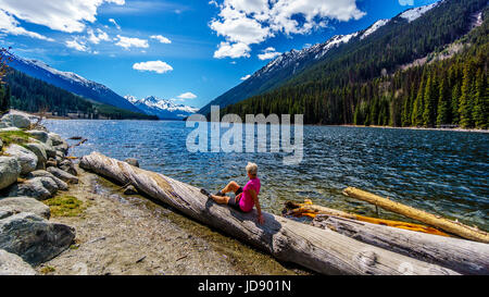Ältere Frau sitzen auf einem Baumstamm, genießen den Blick auf den Schnee begrenzt Gipfel rund um Duffey Lake mit Mount Rohr am Südende des Sees. Stockfoto