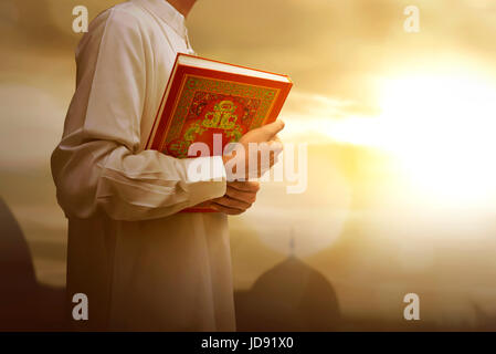 Muslimischen Mann in traditioneller Kleidung, Heilige Buch Koran während Sonnenuntergang Hintergrund hält Stockfoto