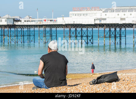 Mann mit einem muslimischen Taqiyah Gebet Mütze, sitzen entlang auf Brighton Beach in der Nähe von Brighton Pier an einem sonnigen Tag. Stockfoto