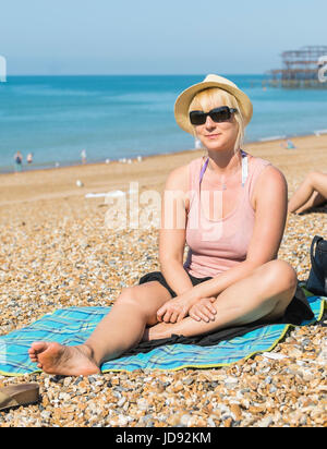 Hübsche junge blonde Frau posiert für ein Foto beim Sitzen auf einem Kiesstrand an einem heißen Tag im Sommer. Stockfoto