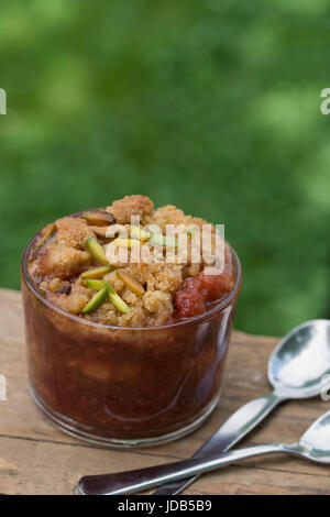 1 Glas Erdbeer-Rhabarber-Crumble, garniert mit Himalajasalz, mit zwei Löffeln auf einem Holztisch in einem grünen Garten. Stockfoto