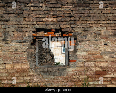 Mauer des alten zerstörten Hauses mit strukturierten Steinen mit einer Öffnung, in der Sie die Fenster eines Hauses Wohn-Dorf sehen können Stockfoto