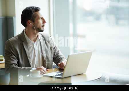 Porträt von reifer nachdenklicher Mann wegsehen, Fenster während mit Laptop im Café mit Kopfhörer Stockfoto
