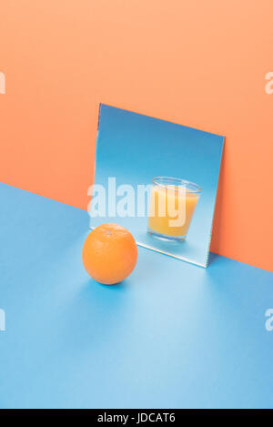 Bild von Orange in der Nähe von Saft in Spiegel auf blaue Tabelle über orange Hintergrund isoliert Stockfoto