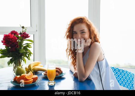 Foto von ziemlich fröhliche junge rothaarige Dame sitzt am Tisch im Haus in der Nähe von Blumen und Früchten. Blick in die Kamera. Stockfoto