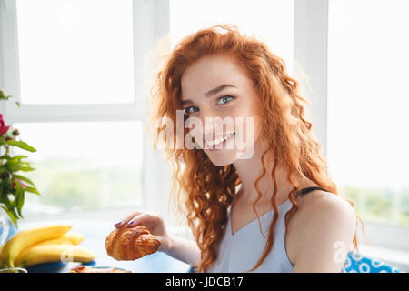 Bild von niedlichen glückliche junge rothaarige Dame sitzt am Tisch im Haus in der Nähe von Blumen und Früchten essen Croissant. Blick in die Kamera. Stockfoto