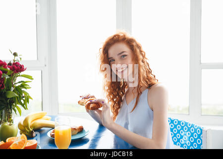 Bild von niedlichen glückliche junge rothaarige Dame sitzt am Tisch im Haus in der Nähe von Blumen und Früchten essen Croissant. Blick in die Kamera. Stockfoto