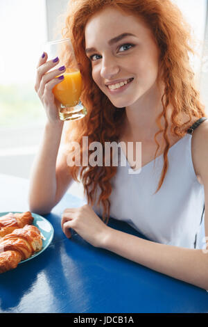 Bild des lächelnden Rothaarige Fräulein am Tisch drinnen Saft zu trinken. Blick in die Kamera. Stockfoto