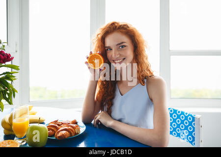 Bild des lächelnden Rothaarige Fräulein am Tisch drinnen halten Orange. Blick in die Kamera. Stockfoto