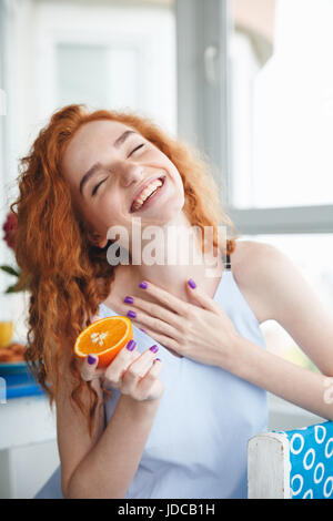 Bild der niedliche junge fröhliche Rothaarige Dame sitzt am Tisch im Haus in der Nähe von Blumen Orange hält. Geschlossenen Augen. Stockfoto