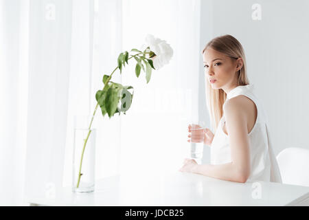 Bild von Ernst blonde Dame drinnen sitzen in der Nähe von Blumen gekleidet in weißen Kleid. Blick in die Kamera. Stockfoto