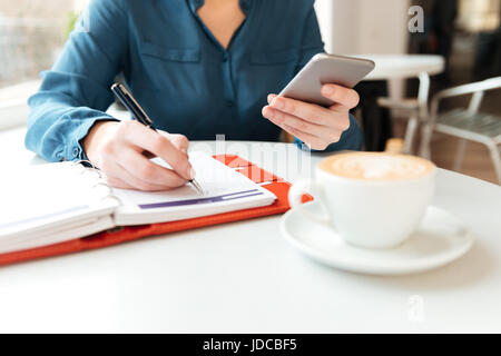 Bild einer Frau am Cafétisch halten Handys und Notizen im Editor abgeschnitten Stockfoto