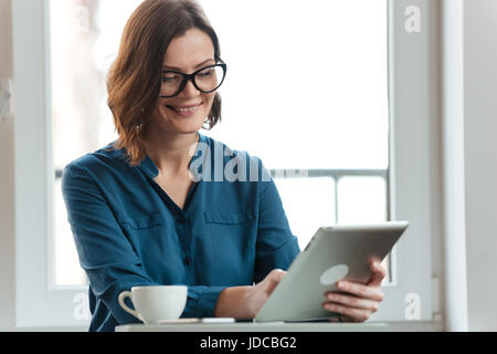 Lächelnde attraktive Frau in Brillen mit Tablet-PC am Cafétisch sitzen und Kaffee trinken Stockfoto