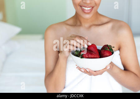 Foto der fröhliche junge afrikanische Frau gekleidet in Handtuch sitzen zu Hause während des Essens Erdbeere beschnitten. Stockfoto