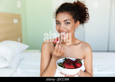 Bild der glückliche junge afrikanische Frau gekleidet in Handtuch zu Hause sitzen und Blick in die Kamera während des Essens Erdbeere. Stockfoto