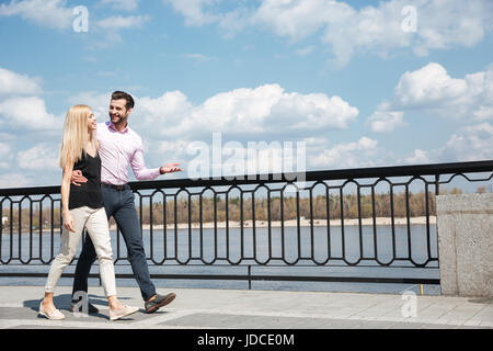 Full-length Schuss von hübschen Liebhaber, fröhlicher Mensch zuversichtlich und hübsche blonde Frau, zu Fuß auf der Brücke in der Nähe Fluss Stockfoto