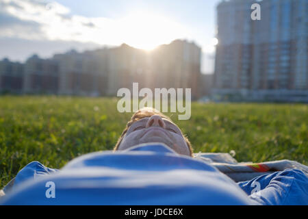 Kaukasische glücklich auf dem grünen Rasen liegend mit ausgestreckten Armen. Stadt in Hintergrundansicht Stockfoto