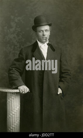 Antike c1910 Fotografie, Studio-Porträt eines Mannes mit Pfeife, Melone und Mantel. Die seltsame Augen hat, und mehrere Ringe trägt. QUELLE: ORIGINALFOTO. Stockfoto