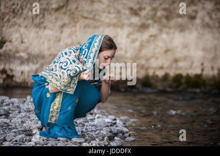 Das Mädchen in die indischen Sari sitzt an dem kleinen Fluss und trinkt aus Händen Stockfoto