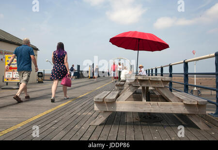 Holz- Picknick Tische & Stühle mit roten Sonnenschirme in Southport, Merseyside, UK. UK Wetter. 20 Juni, 2017. Leichte Winde und lückenhafte Wolken auf Southport Meer als pier Kinderwagen die Meeresbrise aus der Irischen See genießen. Vereinzelt Wolken geben etwas Entlastung von den hohen Temperaturen, als Urlauber und Touristen die Promenade die längste Eisen Pier in Großbritannien. Stockfoto