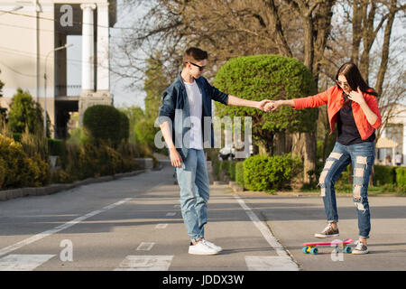 Verliebte Teenager. Der Kerl unterrichtet seine Freundin Skateboard. Datum der Hipster. Er hält ihre Hand. Stockfoto