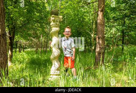 Eine Skulptur in einem grasbewachsenen Wald mit einem kleinen Jungen mit Brille in einem T-Shirt und Shorts an einem Sommertag, Schottland, Großbritannien Stockfoto