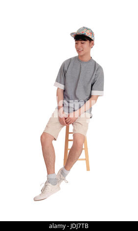 Junge asiatische Teenager sitzen auf einem Stuhl in kurzen Hosen und eine Mütze, isoliert lächelnd, weißen Hintergrund. Stockfoto