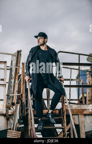 Junger attraktiver Mann im städtischen Hintergrund steht auf einem rostigen Metalltreppe Stockfoto