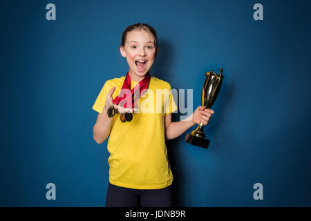 aufgeregt Mädchen mit Medaillen und Trophäen Tasse isoliert auf blau Stockfoto