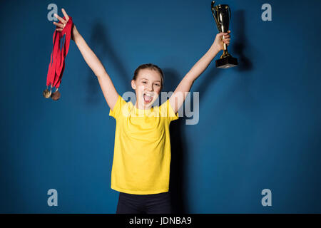 aufgeregt Mädchen mit Medaillen und Trophäen Tasse isoliert auf blau Stockfoto