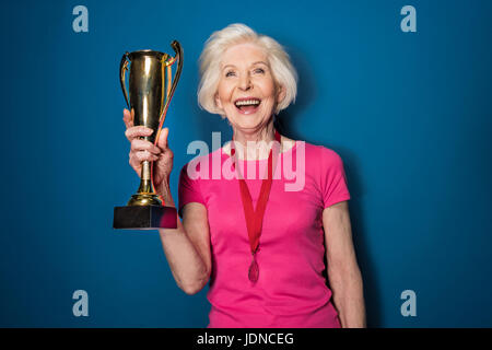 Aufgeregt senior Frau Sportswear halten Trophäe isoliert auf blau Stockfoto