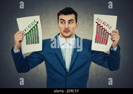 Überrascht junger Geschäftsmann hält zwei Papiere und Blick nach rechts auf die positive Infografik. Erschrocken Gesichts Emotion als Reaktion auf die verschiedenen in Stockfoto