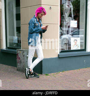 Leeuwarden, Niederlande, 11. Juni 2017: Mädchen mit rosa Haaren lehnt sich an Shop mit Mode für Frauen während der Suche auf dem Handy Stockfoto