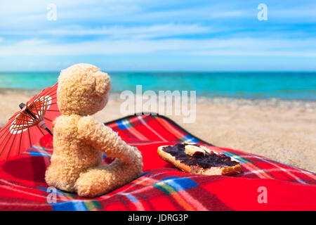 Teddybär mit Decke am Strand sitzen, Marmelade Sonnenschirm und süße Brötchen zum Frühstück Stockfoto