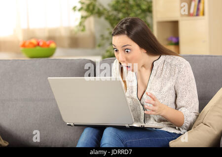 Erstaunt Frau liest auf Linie News in einem Laptop sitzt auf einer Couch im Wohnzimmer in einem Haus-Interieur Stockfoto