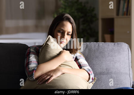 Vorderansicht des ein trauriges Mädchen umarmen eine Kissen sitzen auf einer Couch im Wohnzimmer zu Hause mit einem dunklen Licht im Hintergrund Stockfoto