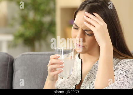 Frau leiden Kopfschmerzen mit einem Wasser-Glas sitzt auf einem Sofa im Wohnzimmer zu Hause Stockfoto