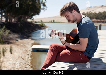 Junger Mann sitzt am Pier, die Gitarre zu spielen Stockfoto