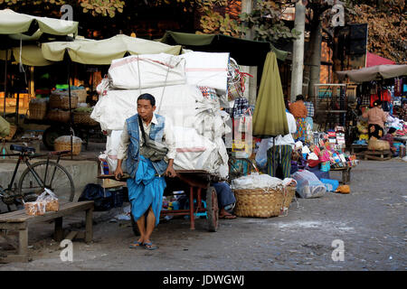 Zegyo Markt/Mandalay - Myanmar 22. Januar 2016: ein Mann mit einem leuchtend blauen Sarong zieht einen großen Wagen mit Körben entlang der Straße. Stockfoto