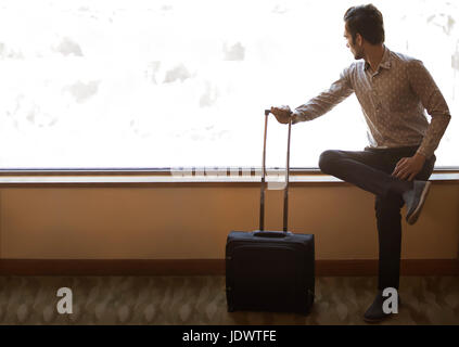 Junger Mann sitzt in der Nähe von Fenster halten Trolley-Tasche Stockfoto