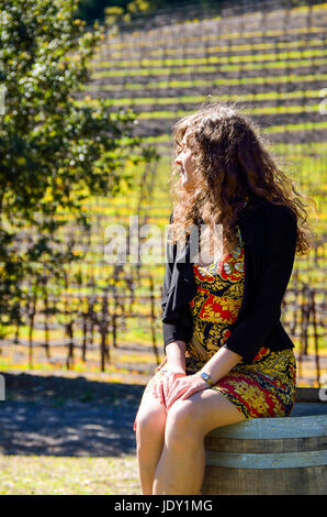 Junge Frau im Kleid sitzt auf Weinfass in Weinberg an sonnigen Tag im Frühling Stockfoto