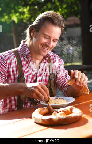 Attraktiver kaukasischer Mann isst traditionelle Käse mit Brezel in einem bayerischen Biergarten Stockfoto