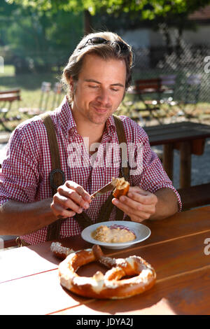 Attraktiver kaukasischer Mann isst traditionelle Käse mit Brezel in einem bayerischen Biergarten Stockfoto