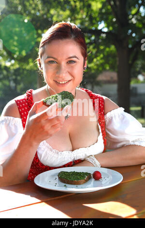 Rote Kopf Frau in typisch bayrischer Tracht isst Brot mit Schnittlauch in einem bayerischen Biergarten Stockfoto