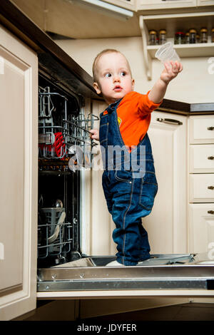 Niedliche Baby stehend auf Geschirrspüler in der Küche und hilft es entladen Stockfoto