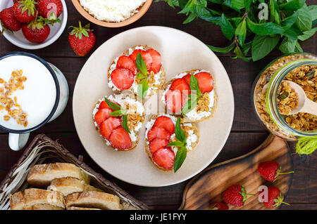 Mini-Sandwiches mit Cottage Käse, frische Erdbeeren, dekoriert mit Minze auf Roggenbrot auf einem dunklen Hintergrund aus Holz. Ansicht von oben. Richtige Ernährun Stockfoto