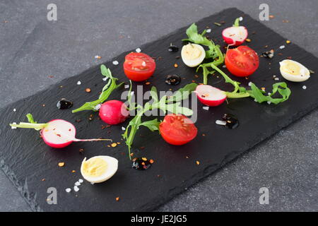 Frische Radieschen, Kirschtomaten, Wachtelei, Parmeggiano mit Balsamico-Glasur auf einem schwarzen Steinplatte. Mediterranes Lebensgefühl. Gesunde Ernährung Stockfoto