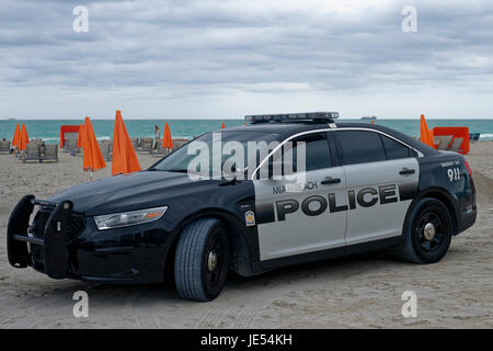 Ein Polizeiauto von Miami Beach am Strand von Miami Beach. Leute liegen an den Strandsitzen. Die orangefarbenen Sonnenschirme sind ein großer Kontrast zum blauen Meer am Horizont sind eine Yacht und ein Frachtschiff sichtbar. Stockfoto
